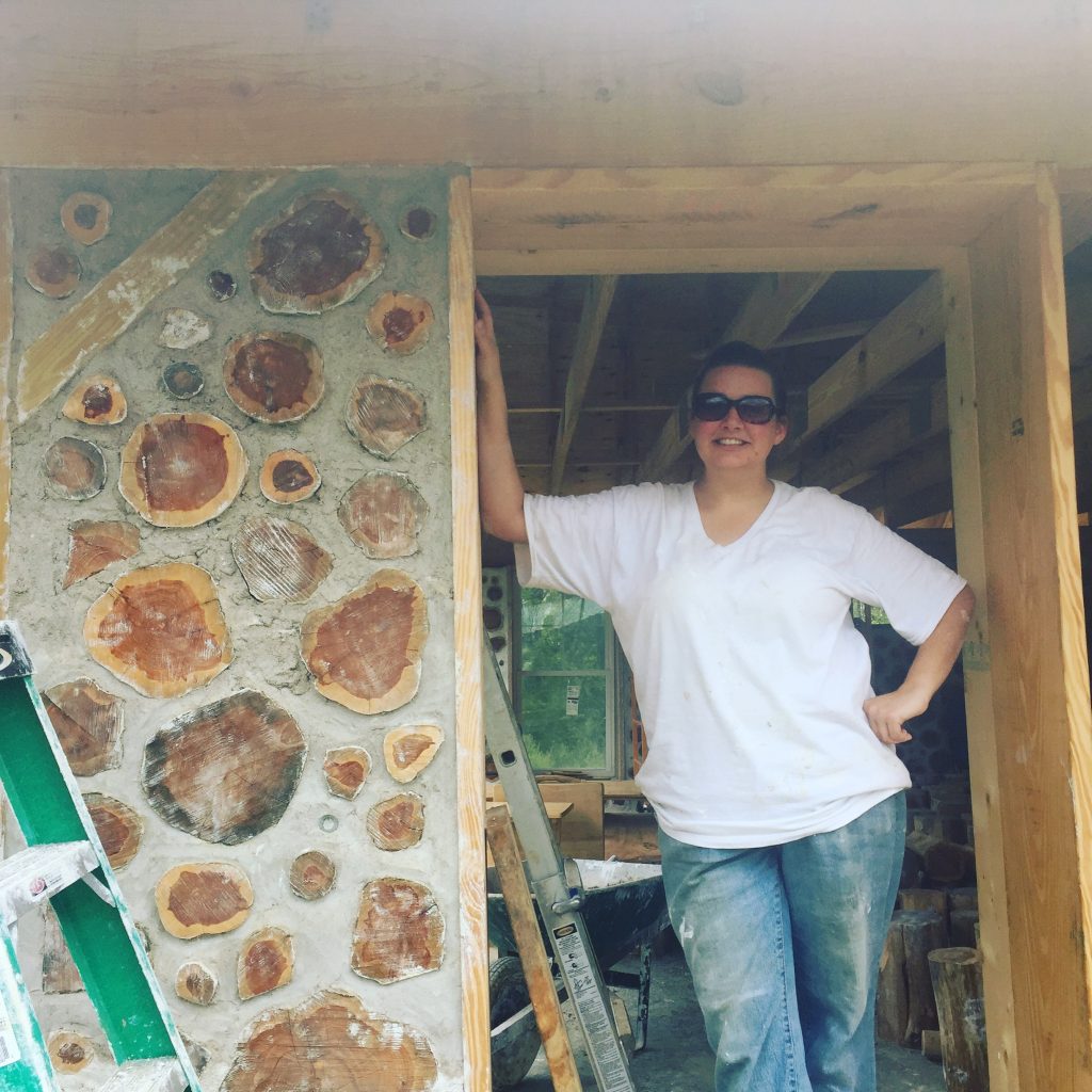 woman standing in a window frame next to cordwood wall