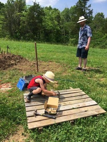 man in blue shirt watching little boy in red shirt and tan hat hammer at a wood pallet
