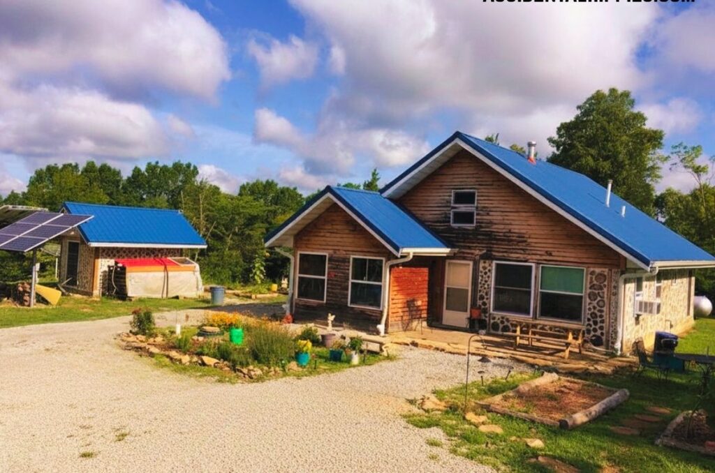 Cordwood House with blue roof next to a similar shed with gravel paths and garden beds and a blue sky with puffy clouds